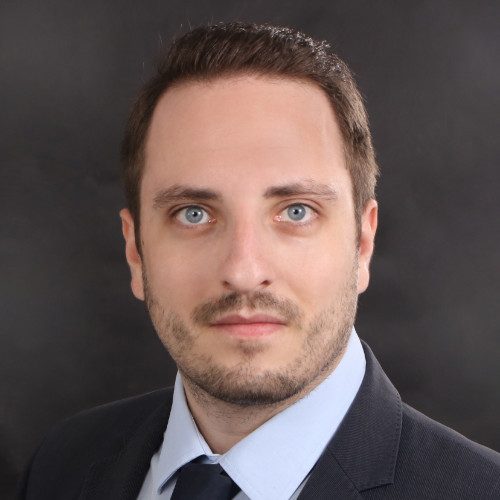 Mann mit kurzem Haar, blauen Augen und Anzug vor einem dunklen Hintergrund.